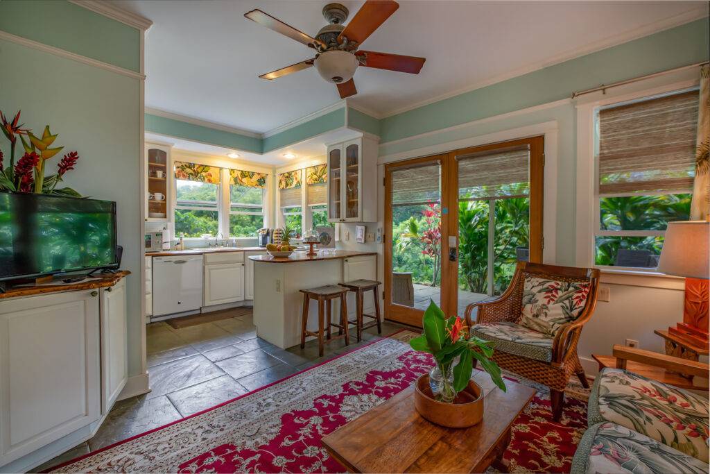 Cottage kitchen with bar, counters, and sink. Double glass doors, a tropical upholstered chair, a tropical settee, a corner table lamp and coffee table. A small tv.