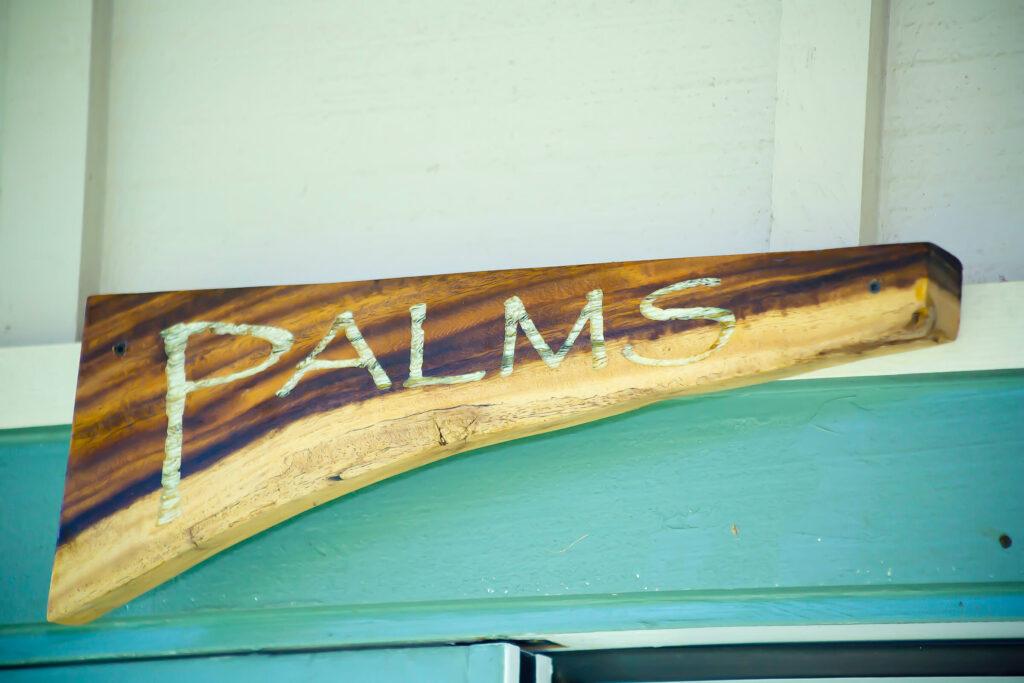 A wooden sign with white letters that reads Palms